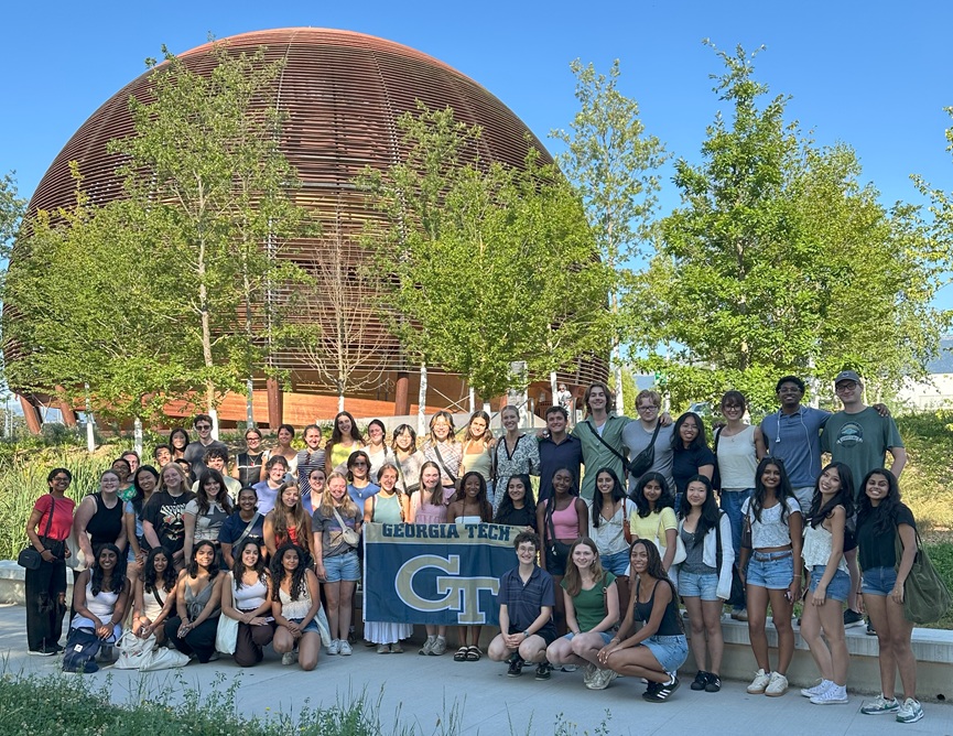 Approximately 50 students from the BEST-Lyon, France Program stand outside of the “Globe of Science and Innovation,” a wooden structure at the entrance of CERN’s visitors center. Some of the students are holding a flag that says Georgia Tech.
