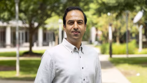 Portrait of an individual standing on a paved campus walkway, wearing a light-colored button-down shirt. Trees, landscaped green spaces, and campus buildings appear in the softly blurred background, with daylight illuminating the outdoor scene. The image is framed from the waist up, with the individual centered in the foreground.